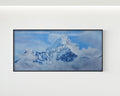 A framed landscape art print depicting a snow-covered mountain peak rising through blue sky and clouds, mounted on a plain white wall.