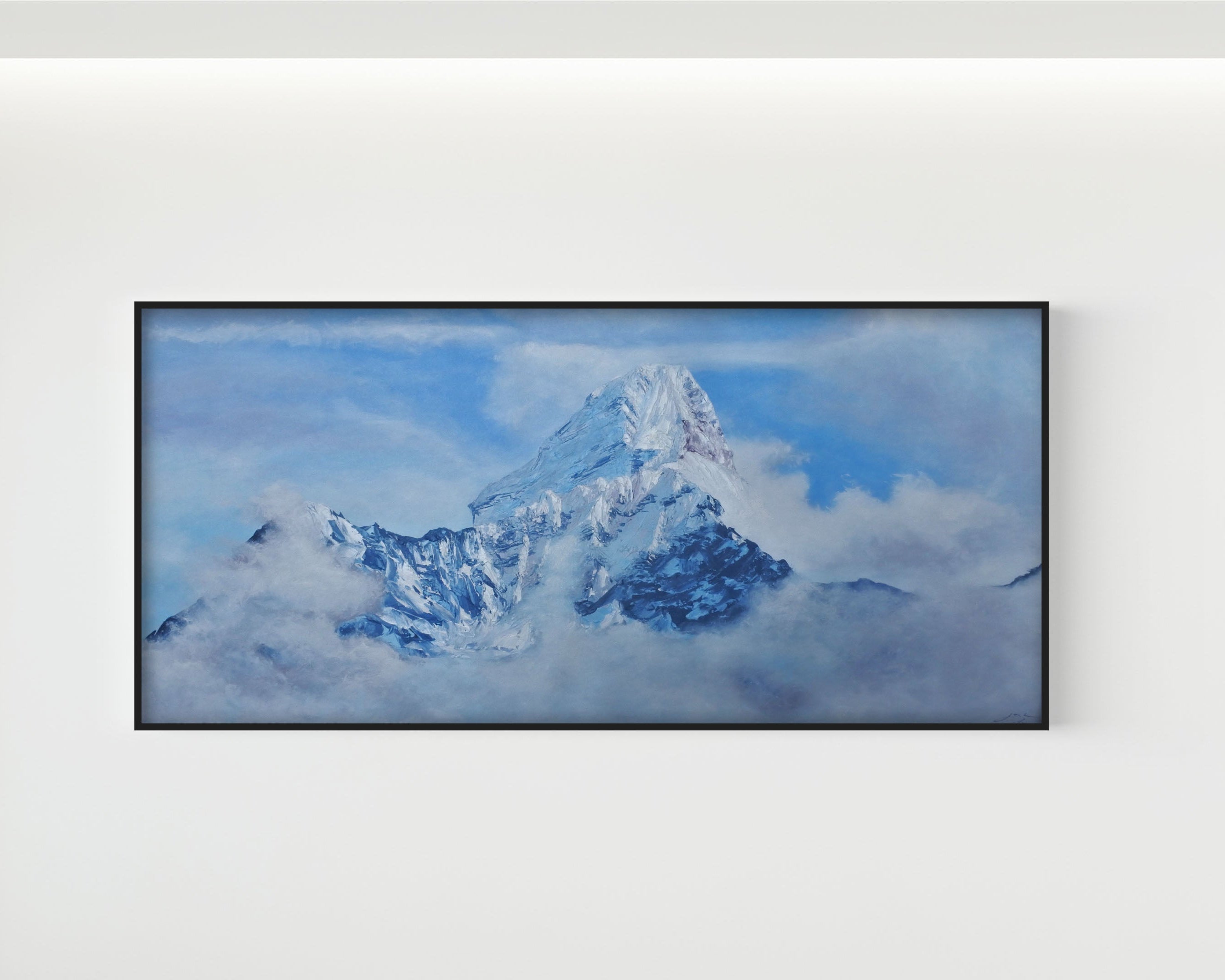 A framed landscape art print depicting a snow-covered mountain peak rising through blue sky and clouds, mounted on a plain white wall.