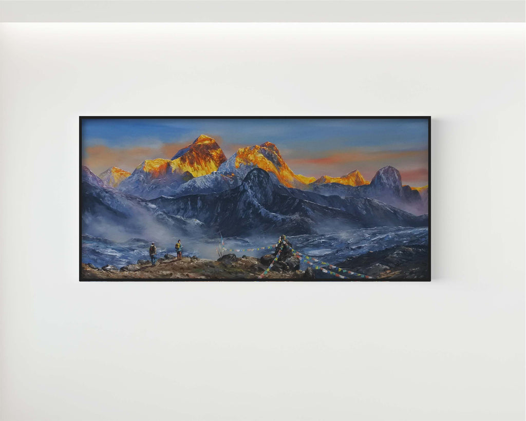 A framed horizontal landscape painting of snow-capped mountain peaks at sunrise, with golden-orange sunlit ridges and deep blue-gray shadows, displayed on a light gray wall.