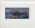 A framed horizontal landscape painting of snow-capped mountain peaks at sunrise, with golden-orange sunlit ridges and deep blue-gray shadows, displayed on a light gray wall.